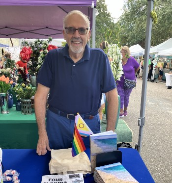 Image of James Williams, an older gentlemen wearing a blue shirt standing by his book stand attending a book fair decorated with a Pride Flag