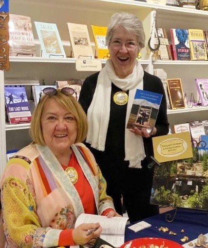 Authors Judilee Butler and GaGa Gabardi at a book signing event. GaGa Gabardi is seated, signing books, while Judilee Butler stands beside her holding one of their co-authored titles.